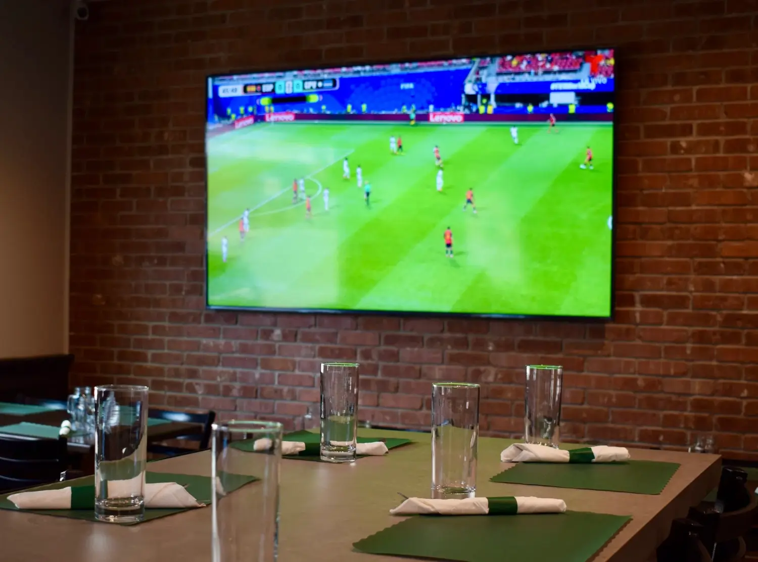 A TV on the brick wall above tables set with green napkins.